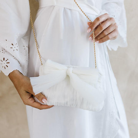 A woman in a white dress holds a stylish white clutch bag with a bow